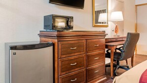 A hotel room with a wooden dresser, a television, a desk, a chair, a refrigerator, and a mirror.