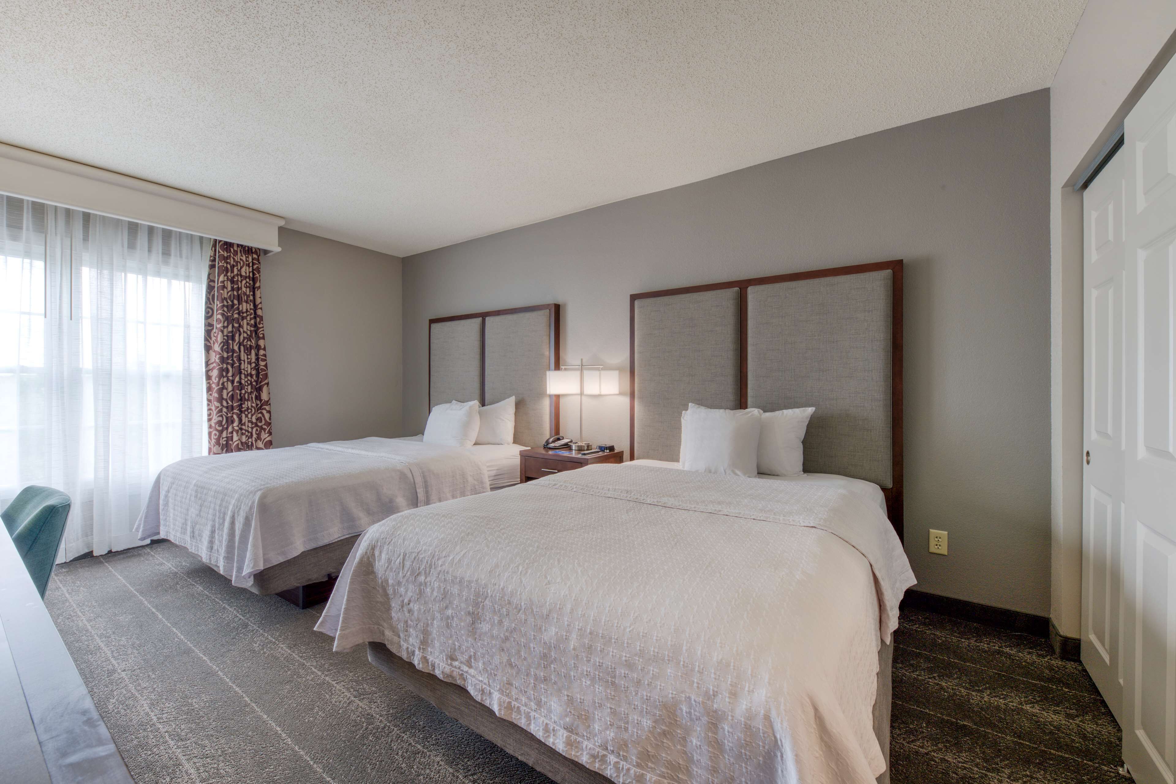 A hotel room with two beds, a nightstand, a lamp, and a window with curtains.