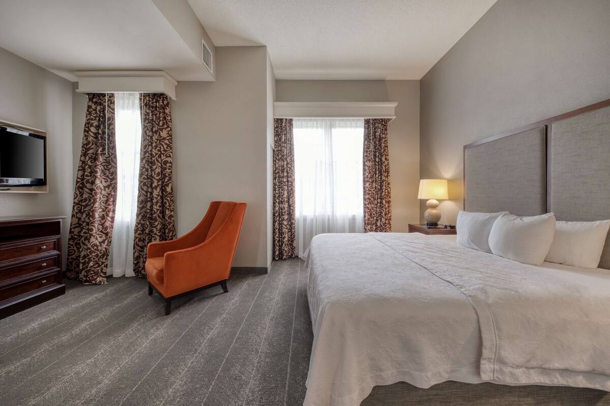 A hotel room with a large bed, a television, an orange chair, and patterned curtains.