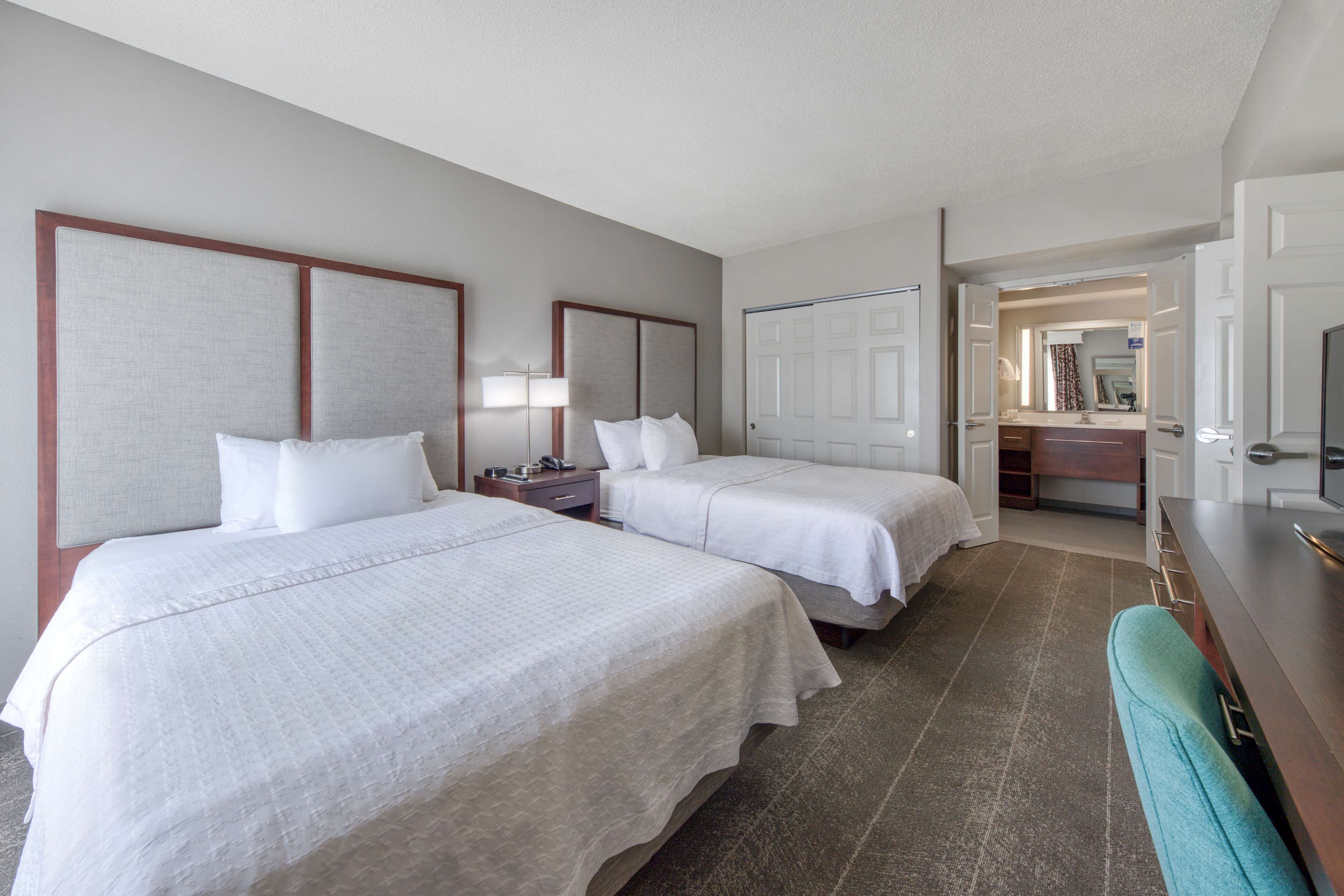 Ein Hotelzimmer mit zwei Betten, einem Schreibtisch, einem Stuhl und einem durchgegangenen Türspalt erkennbaren Badezimmer.