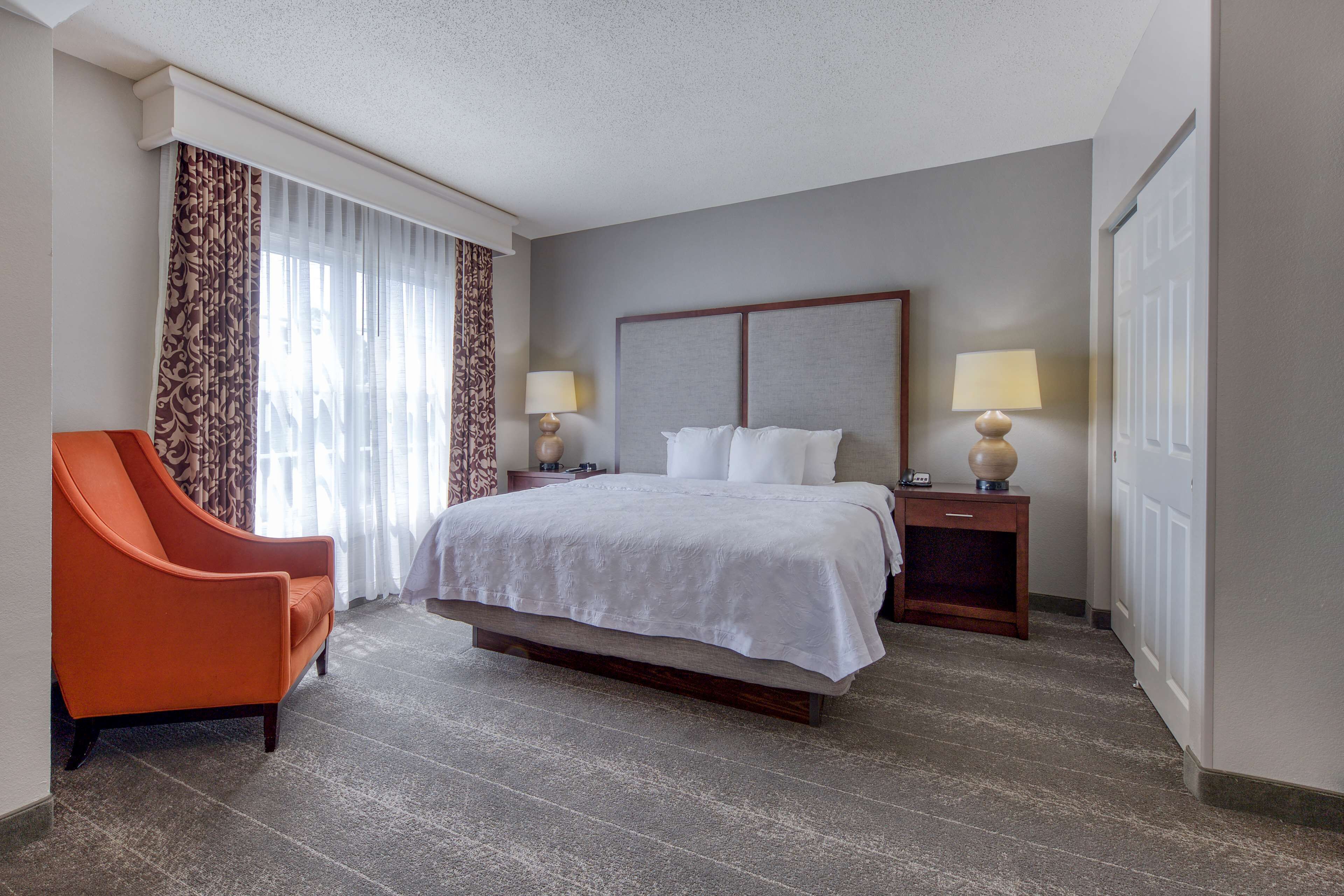 A hotel room with a large bed, two bedside tables with lamps, a wooden nightstand, a curved orange armchair, and a window with patterned curtains.