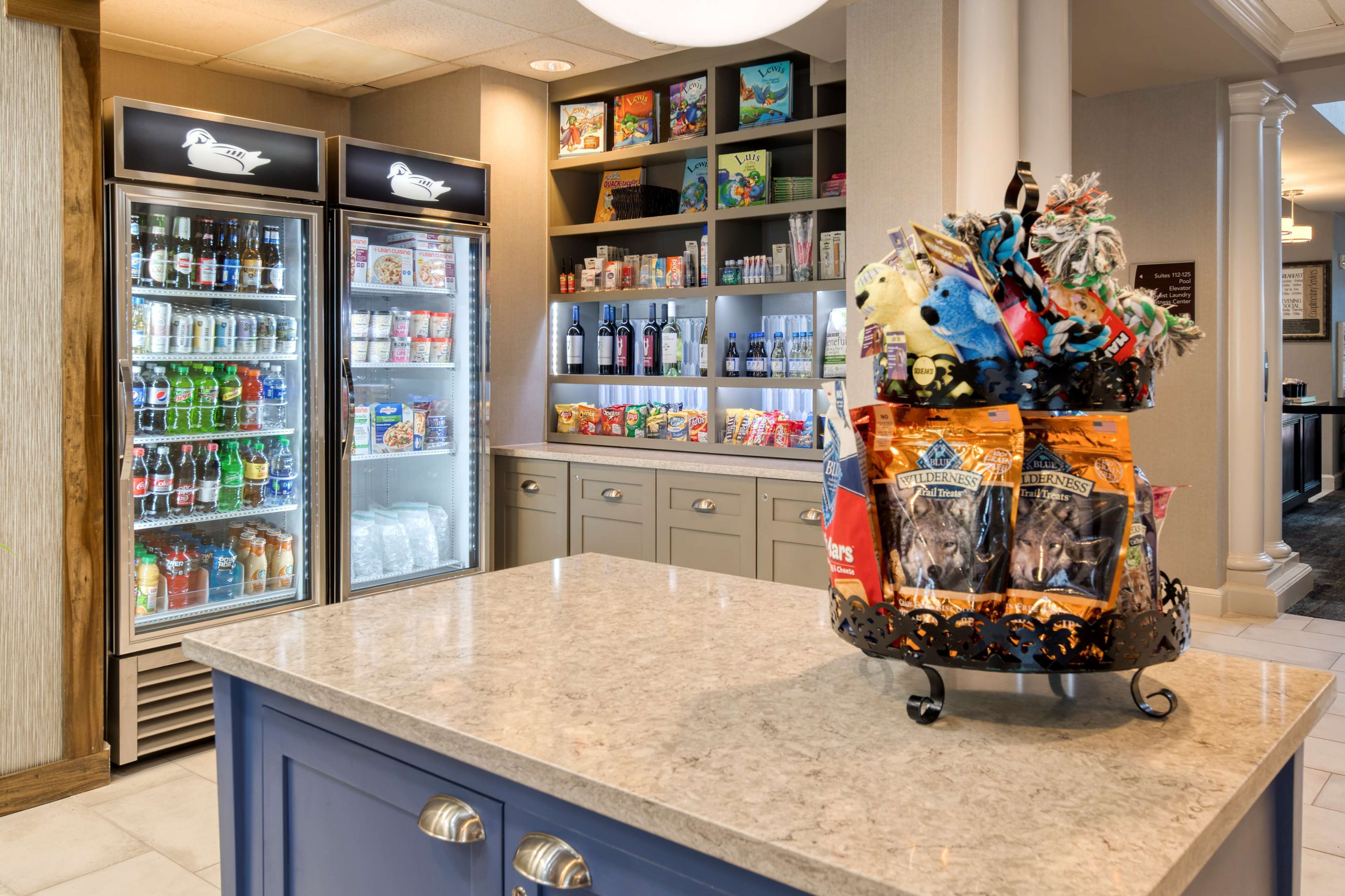 A hotel lobby with a marble counter, refrigerated beverage dispensers, snack shelves, and a decorative gift basket.