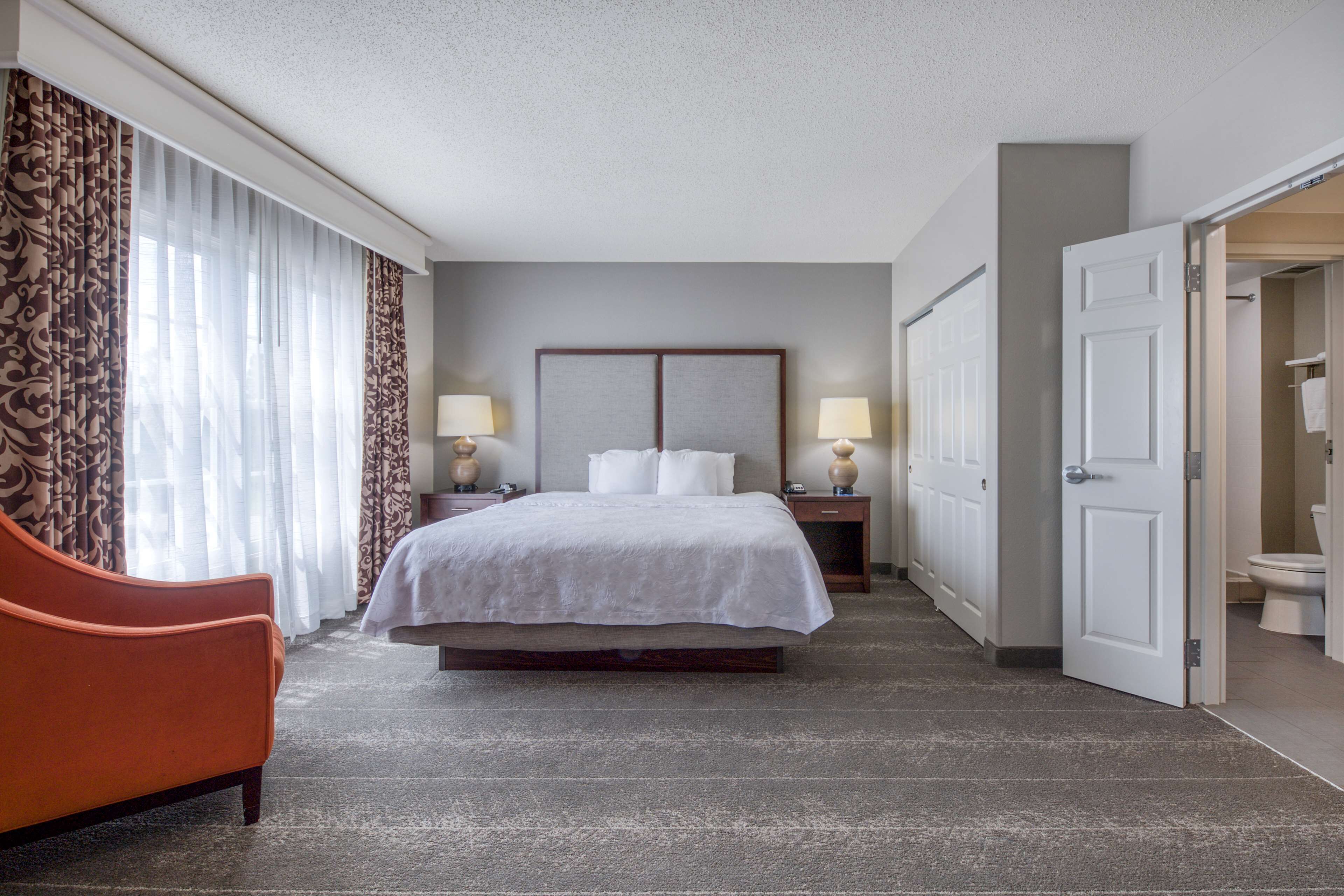 A hotel room with a bed, a nightstand, a lamp, a chair, and a bathroom door.