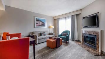 A modern hotel room with a sofa, armchairs, a dining table, a flat-screen TV, and a fireplace.