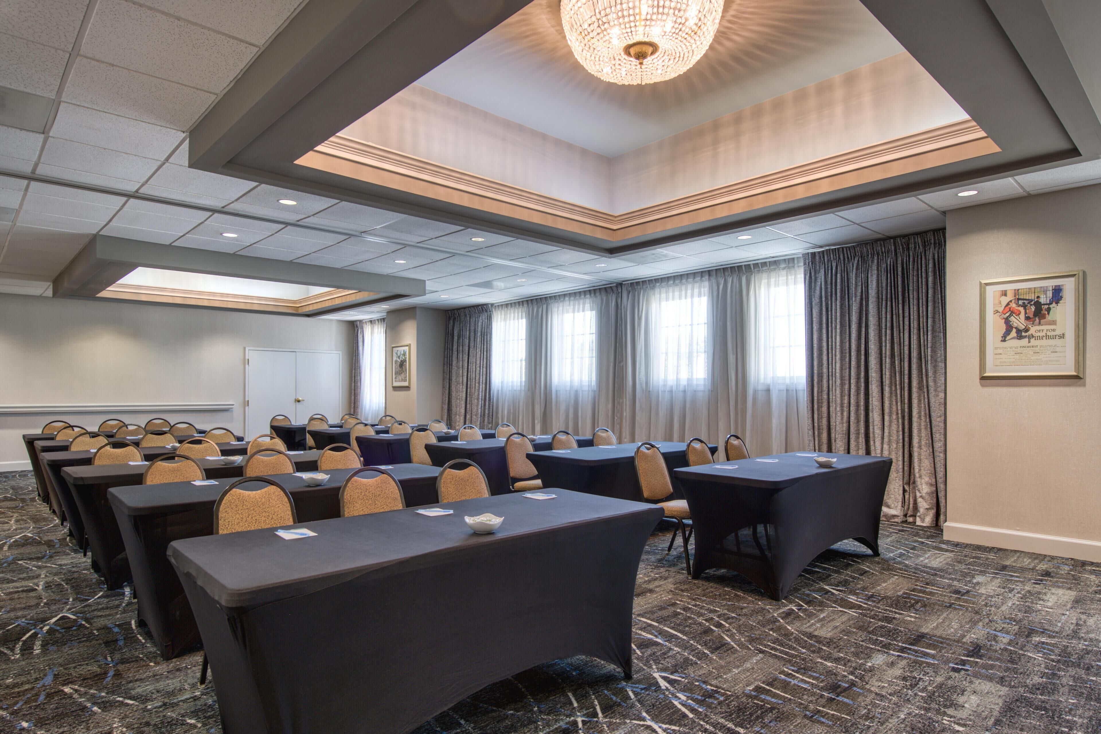 A conference room with multiple tables and chairs, a chandelier, and large windows with curtains.