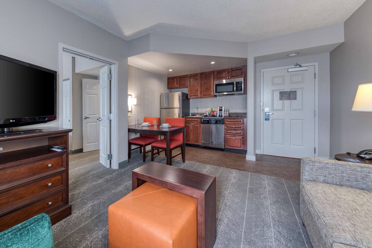 A modern kitchen with stainless steel appliances, a dining area with a red table and chairs, and a living room with a sofa and a TV.