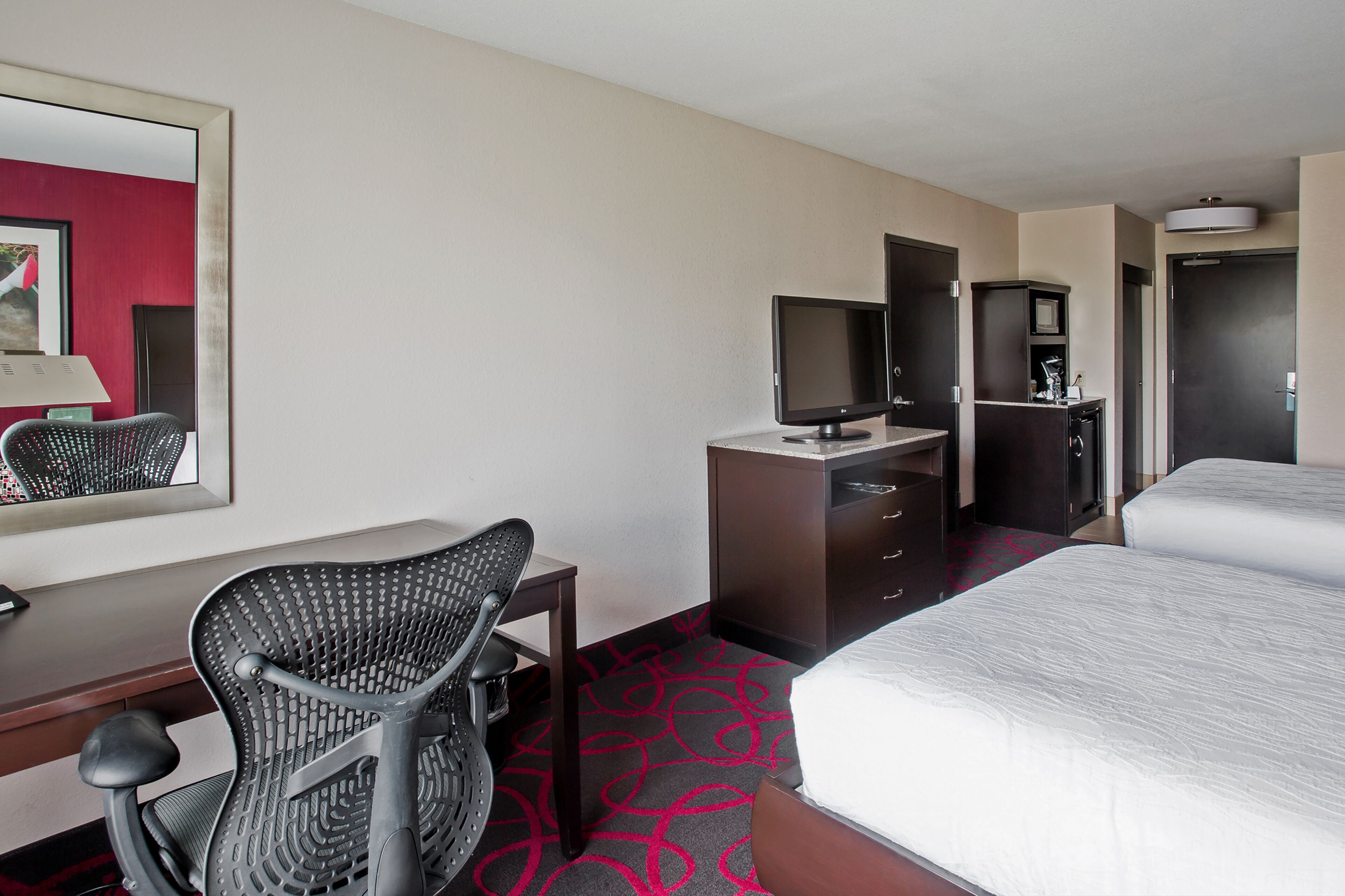 Una habitación de hotel con cama, escritorio con silla, televisor, armario y espejo.