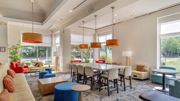 A spacious living area with a large sofa, blue ottoman, and a dining table with chairs. There are hanging pendant lights and a view of greenery outside.