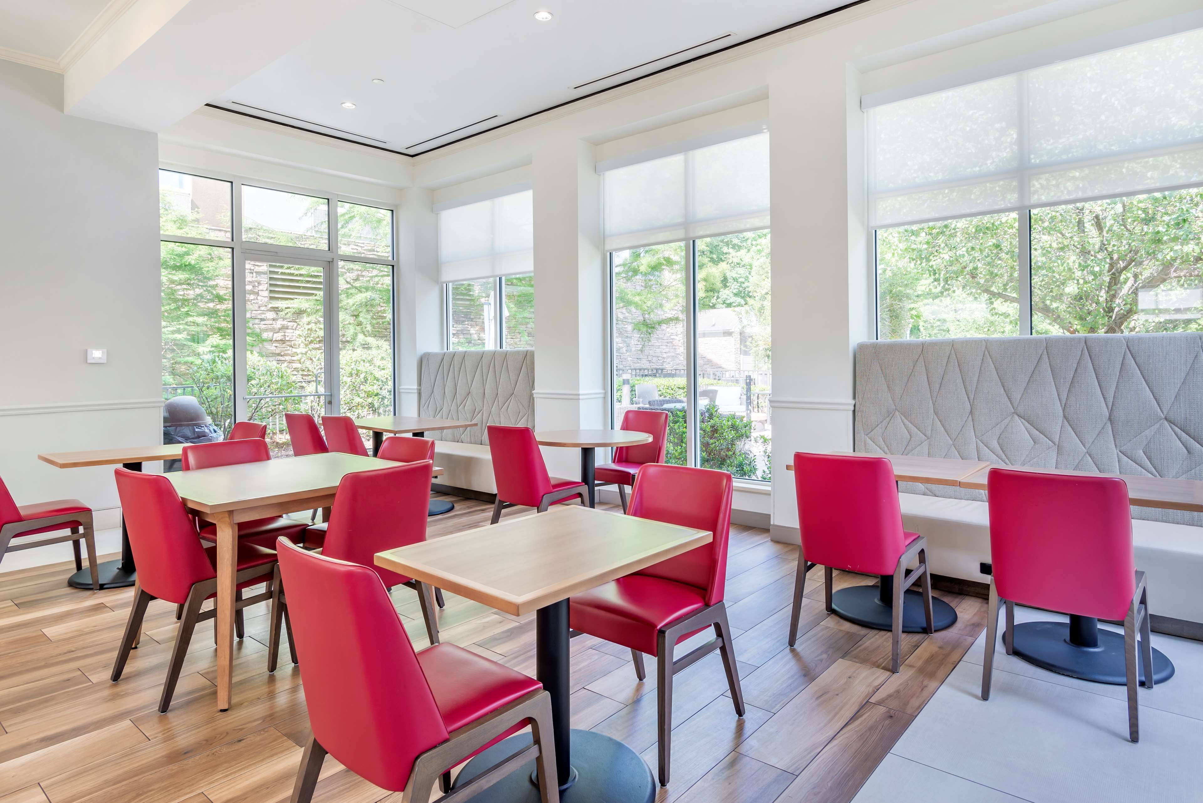 A modern dining area with red chairs and wooden tables, large windows with a view of greenery, and a cushioned bench area.