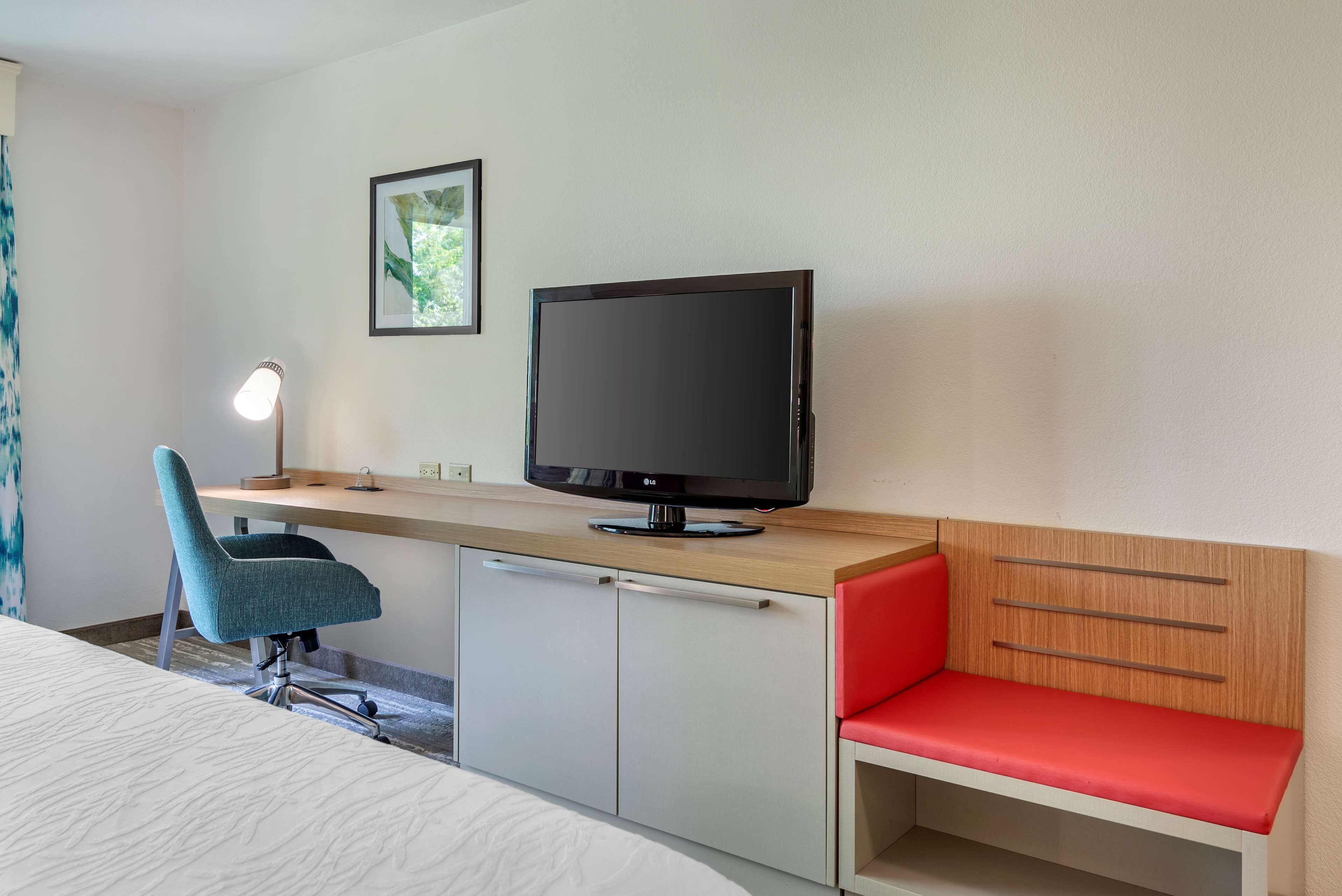 Quarto de hotel com uma cama, uma escrivaninha com televisão, uma cadeira, uma mesa de cabeceira com abajur e um banco vermelho.