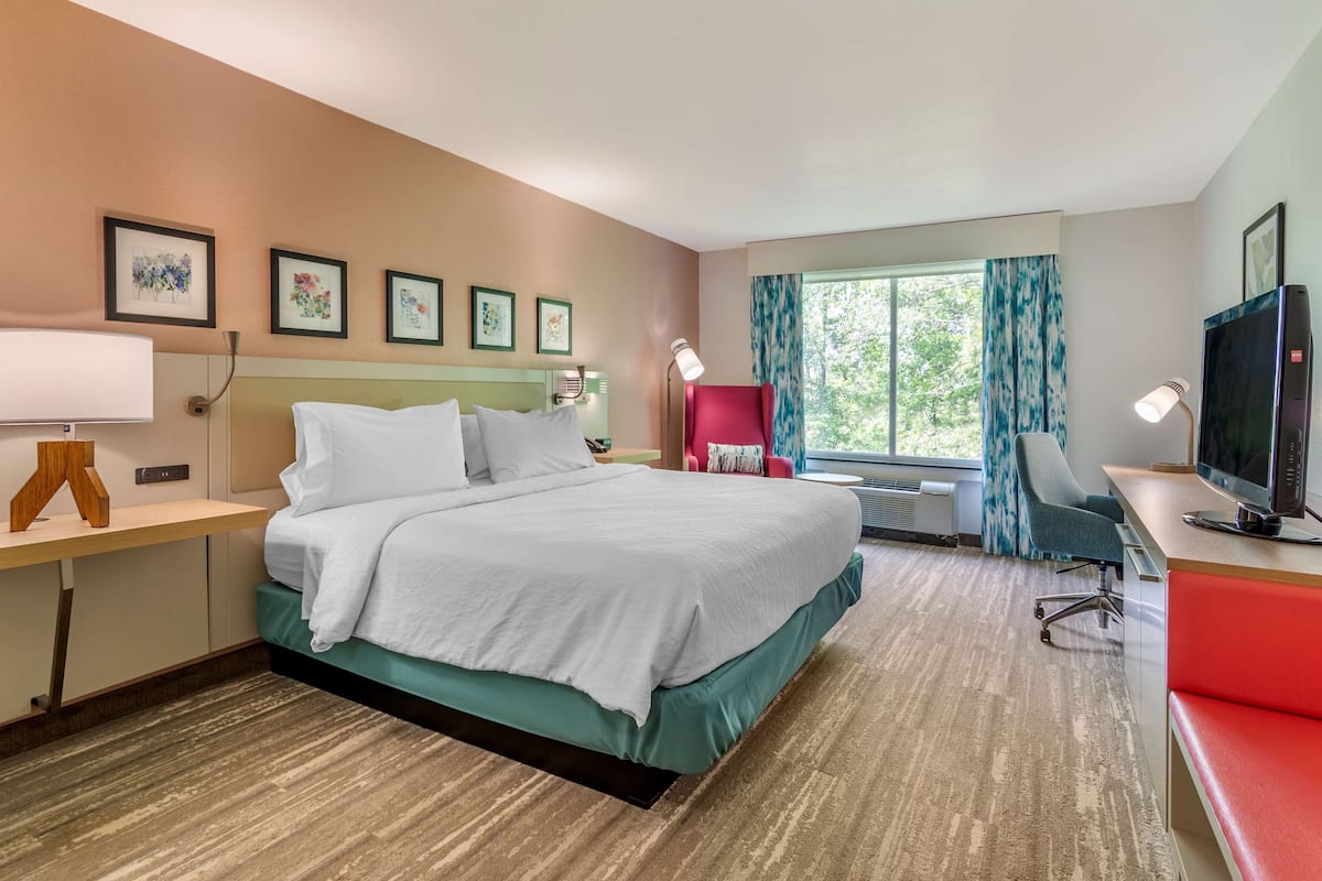 A hotel room with a bed, desk, chair, TV, and large window with curtains.
