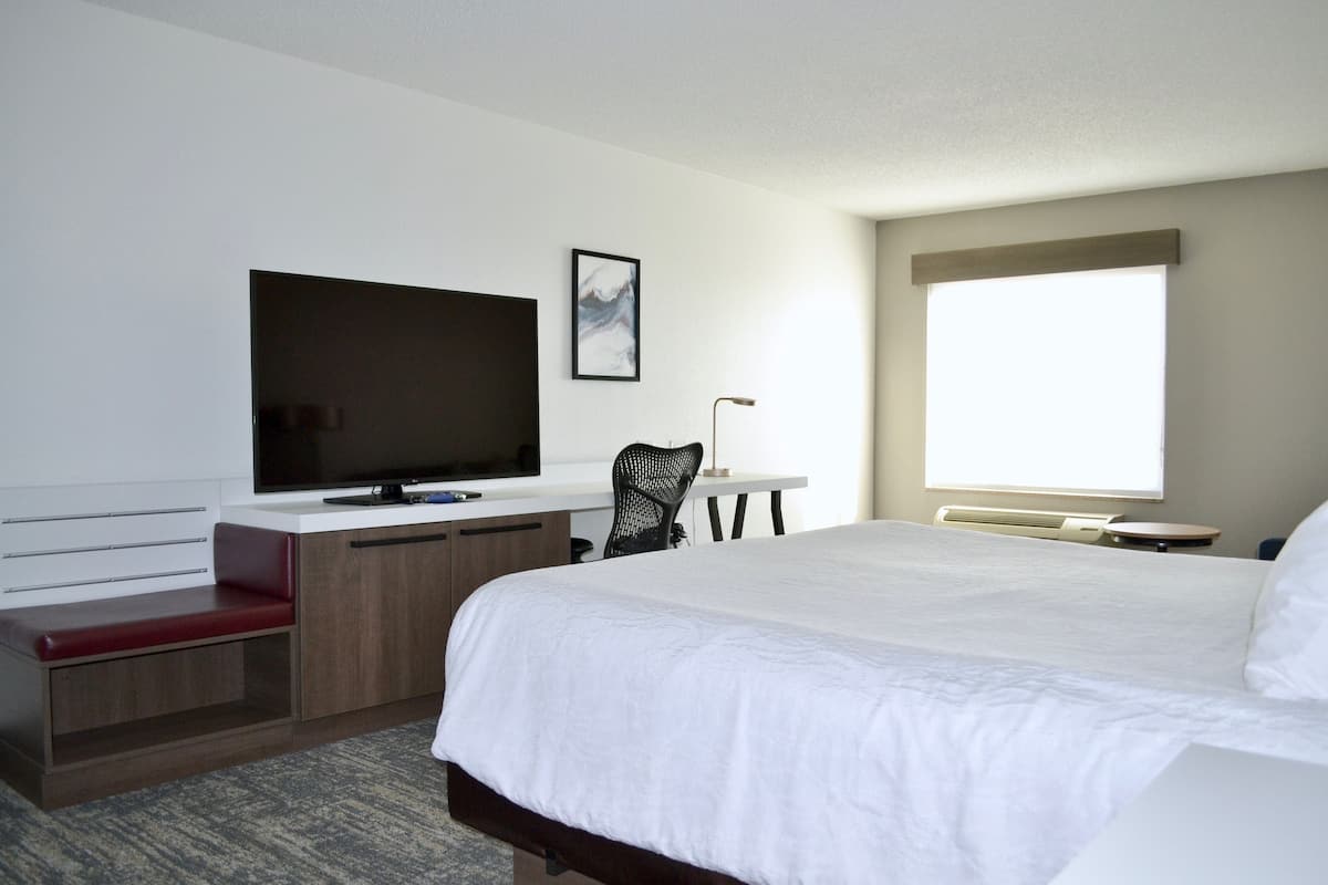 A hotel room with a large bed, a desk with a flat-screen TV, a chair, a window, and a red bench.