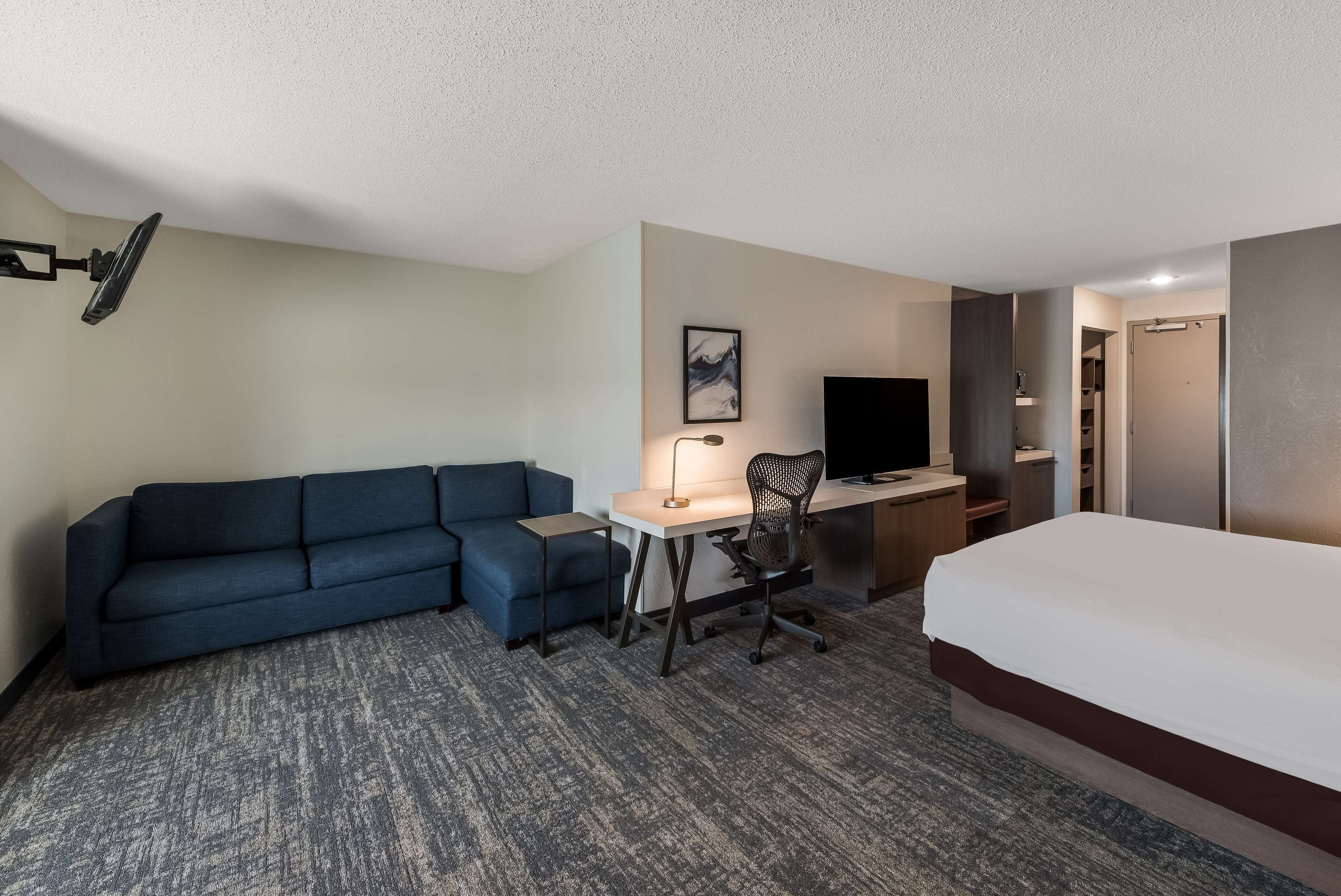 A hotel room with a bed, a desk with a chair, a sofa, a TV, and a wall-mounted lamp.