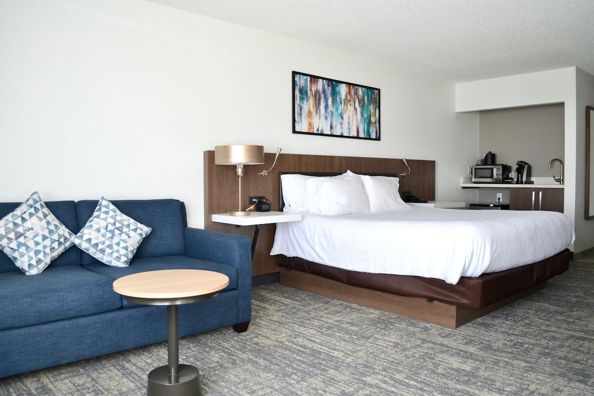 A hotel room with a bed, a sofa, a small table, a lamp, a painting, and a kitchenette.