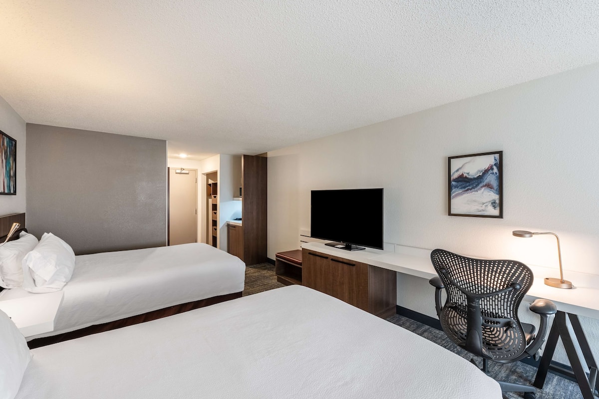 A hotel room with two beds, a desk with a TV, a chair, and a lamp.