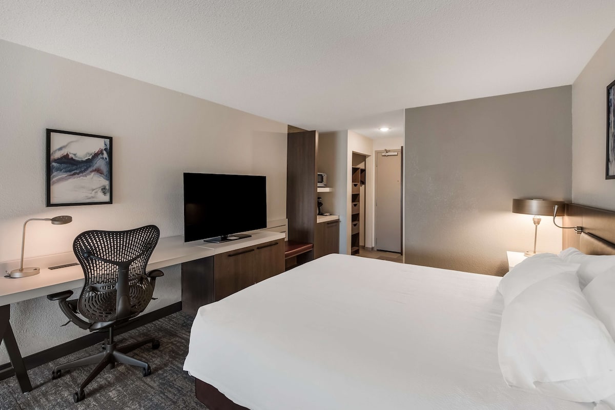 A hotel room with a bed, a desk with a computer, a chair, a TV, and a painting on the wall.