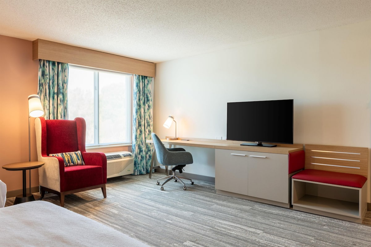 A hotel room with a bed, a desk with a TV, a red armchair, a grey office chair, and a bedside table.