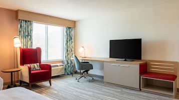 Quarto de hotel com cama, escrivaninha com televisão, poltrona vermelha, cadeira de escritório cinza e mesa de cabeceira.