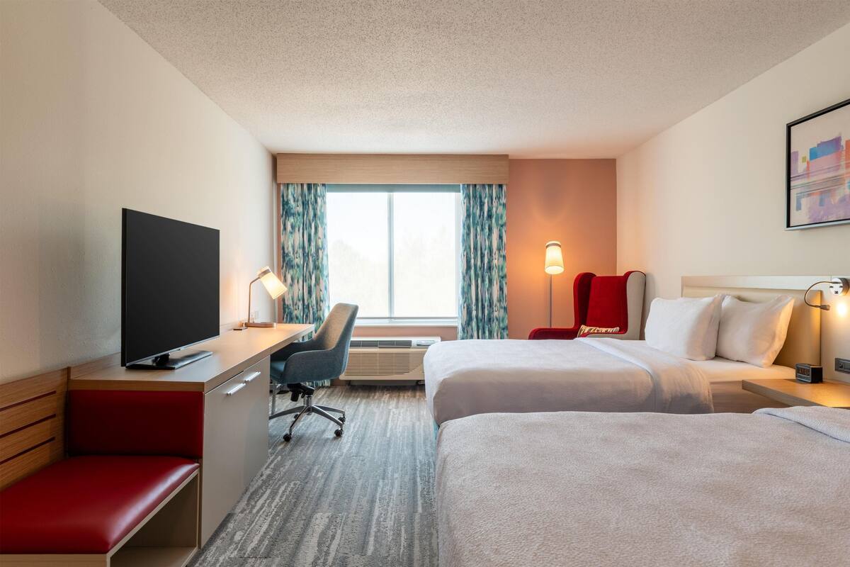 A hotel room with two beds, a desk with a TV, a chair, a lamp, and a window with curtains.