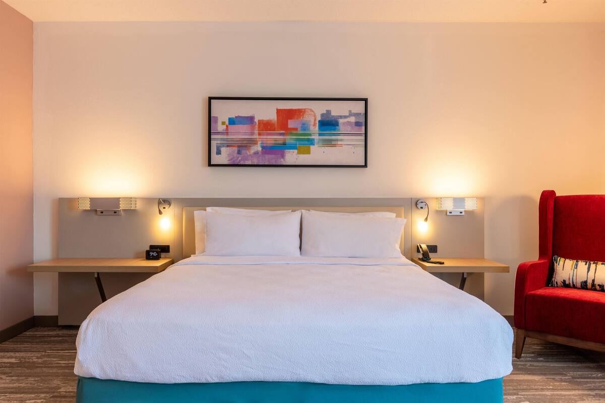 A hotel room with a large bed, two bedside tables, a red chair, and a colorful abstract painting on the wall.
