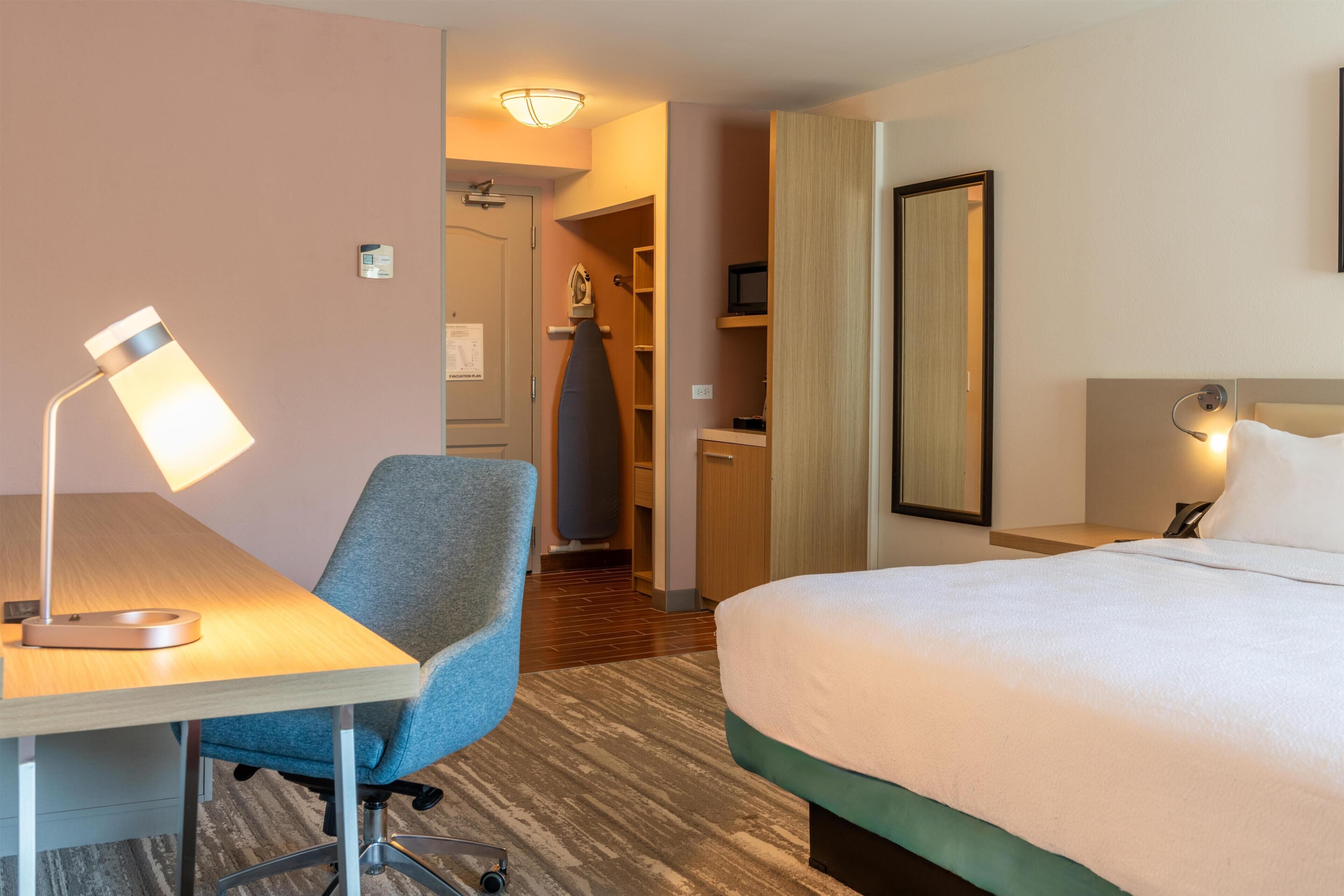 A hotel room with a bed, desk, chair, lamp, and a mirror.