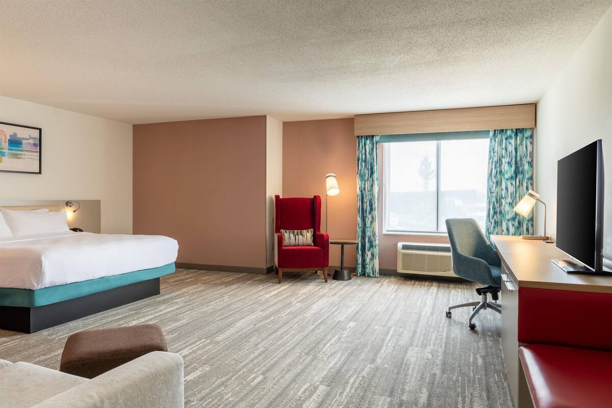 A hotel room with a bed, a desk, a chair, a red armchair, a TV, and a window with curtains.