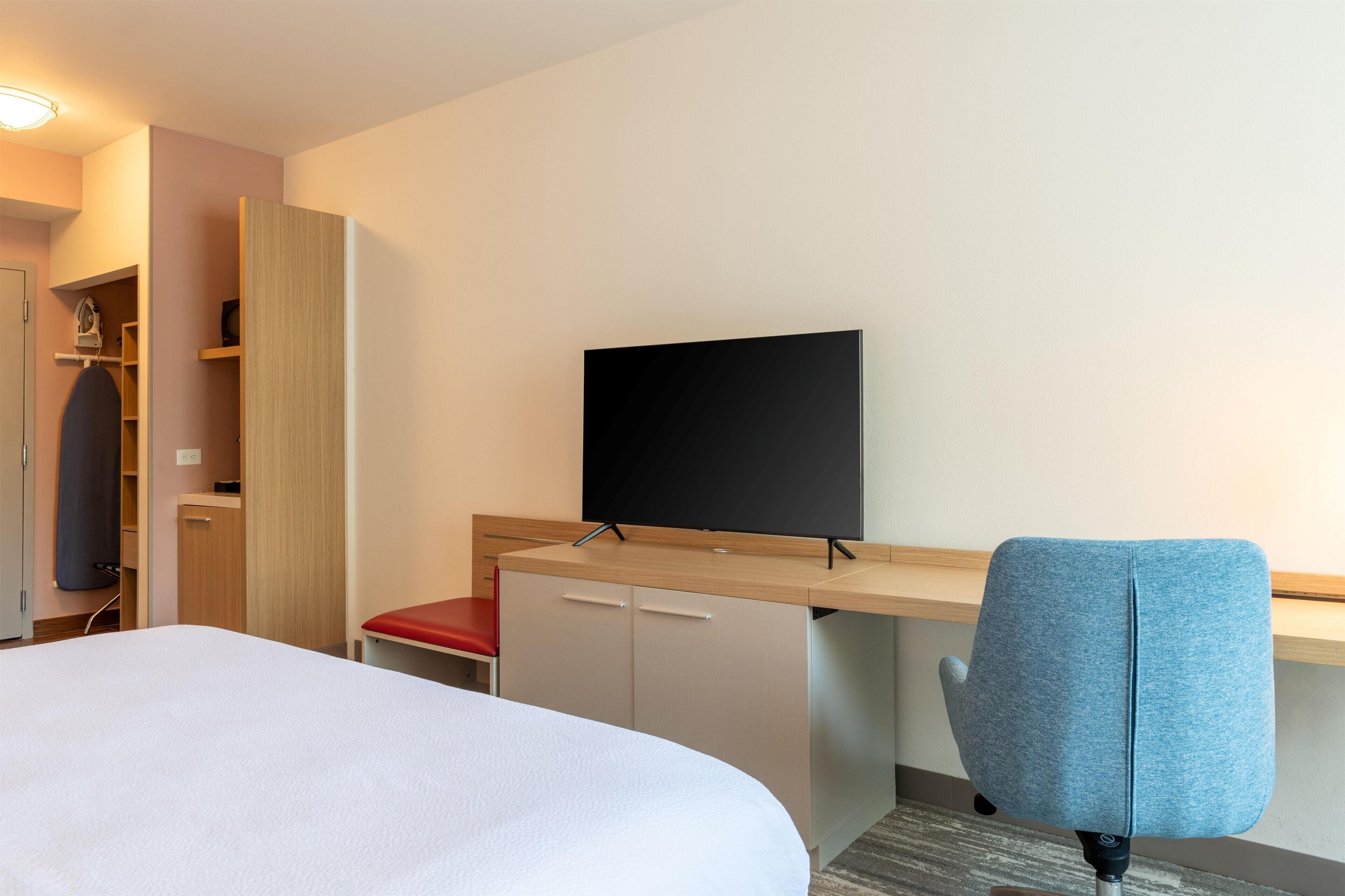 A hotel room with a bed, a desk with a television, a chair, and a red seat.