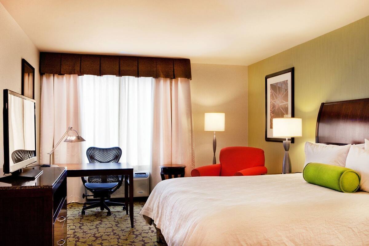 A hotel room with a bed, a desk with a computer, a chair, a red chair, a lamp, and a framed picture on the wall.
