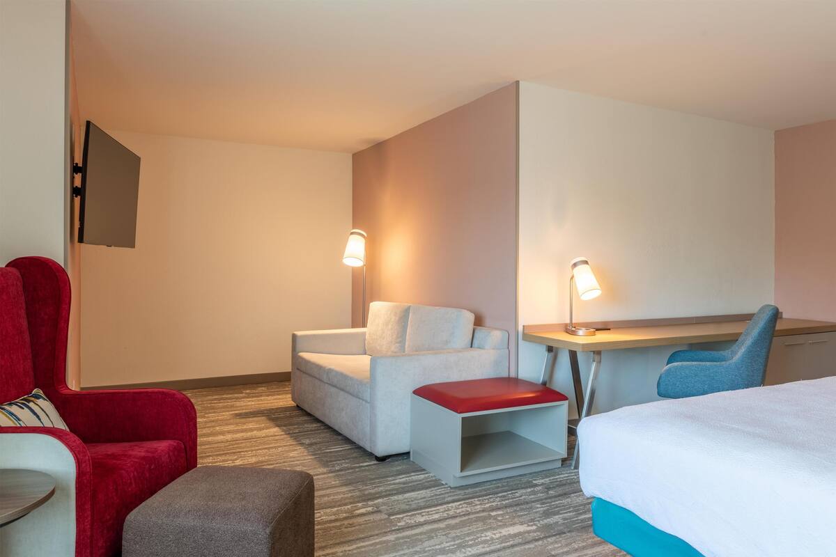 A hotel room with a bed, a desk, a chair, a sofa, and a TV mounted on the wall.