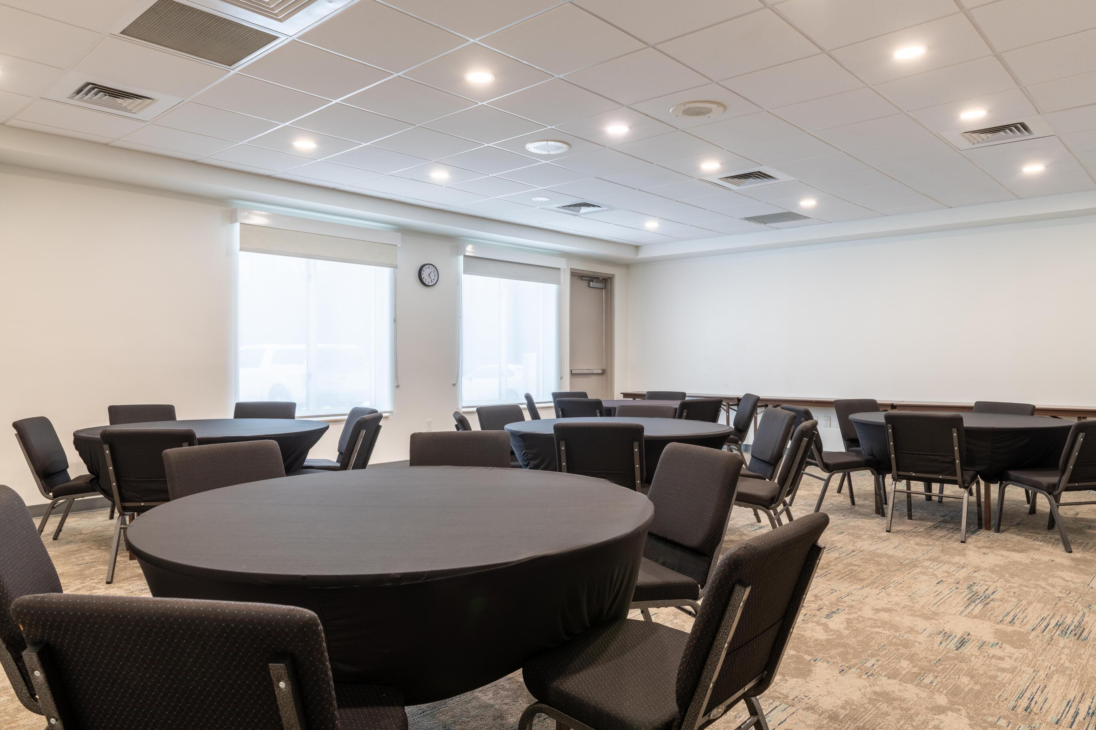 A conference room with multiple tables and chairs, large windows, and a clock on the wall.