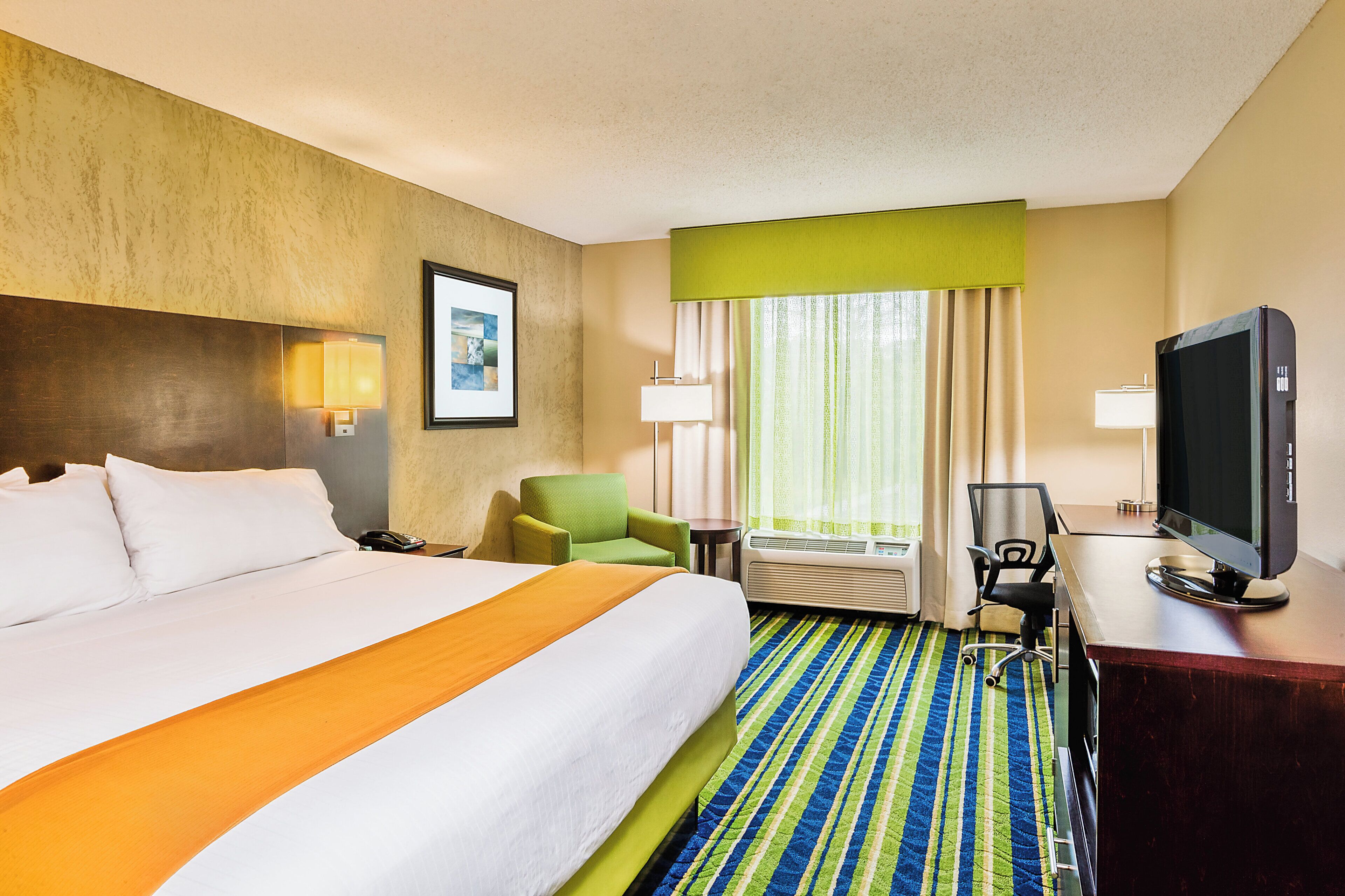 A hotel room with a large bed, a desk with a television, a chair, and a window with curtains.