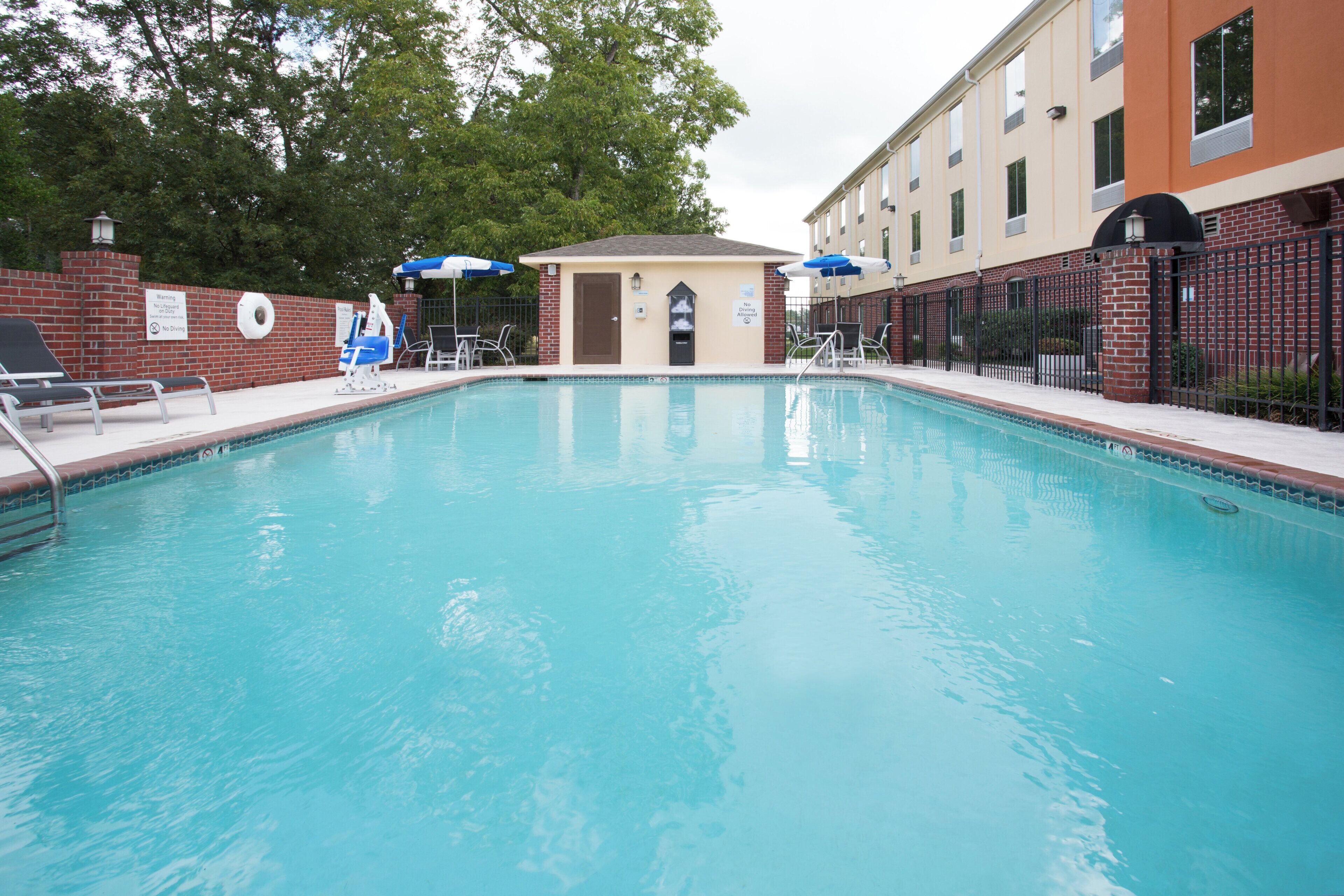 A swimming pool with lounge chairs and umbrellas, a lifeguard stand, and a brick wall with a sign.