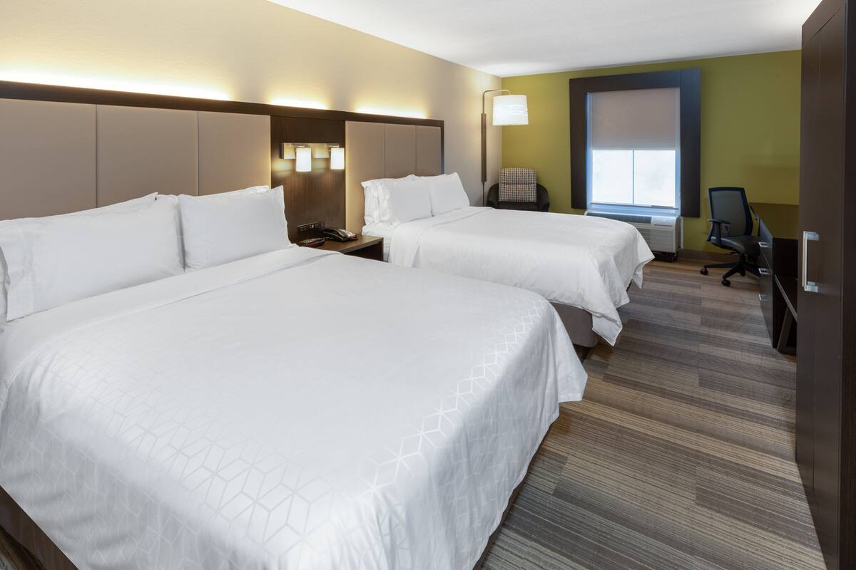 A hotel room with two beds, a desk, a chair, a nightstand, and a window with blinds.