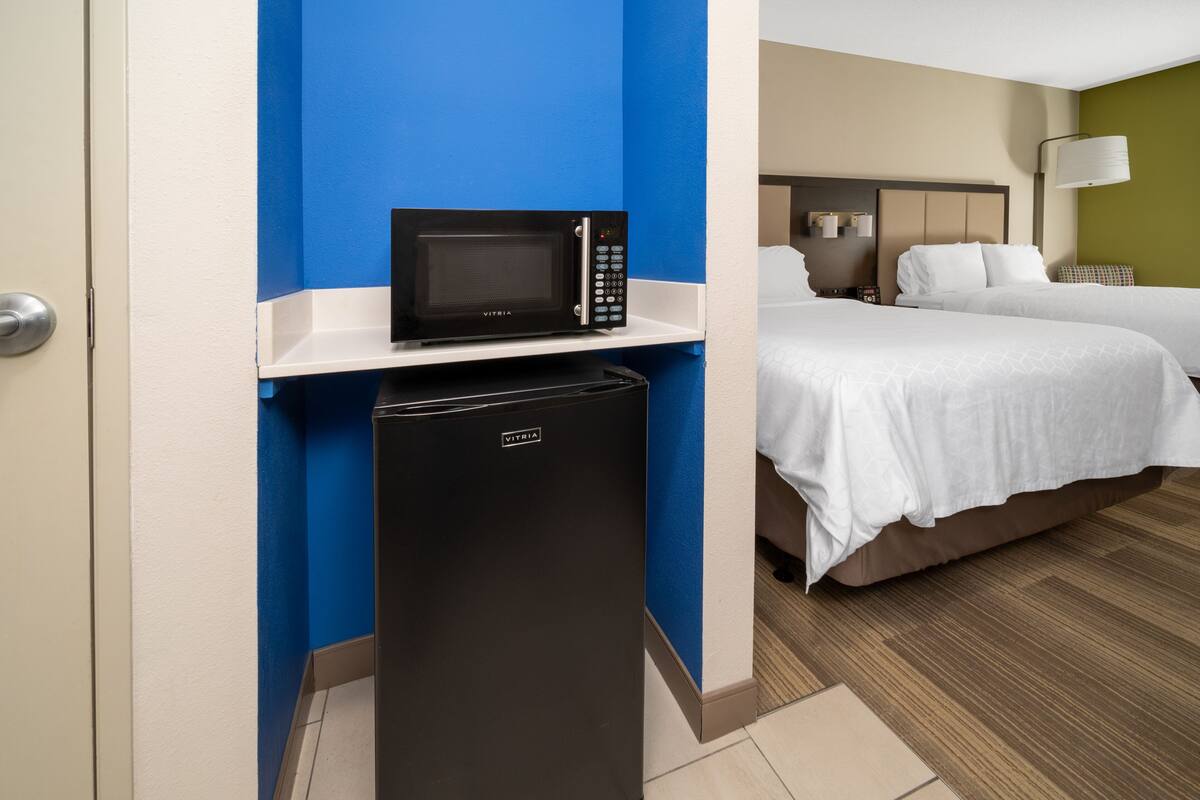 Une chambre d’hôtel de taille réduite, équipée d’un four à micro-ondes posé sur une petite étagère, d’un réfrigérateur compact, de deux lits avec du linge de lit blanc, d’une table de chevet avec une lampe et d’un luminaire fixé au mur.