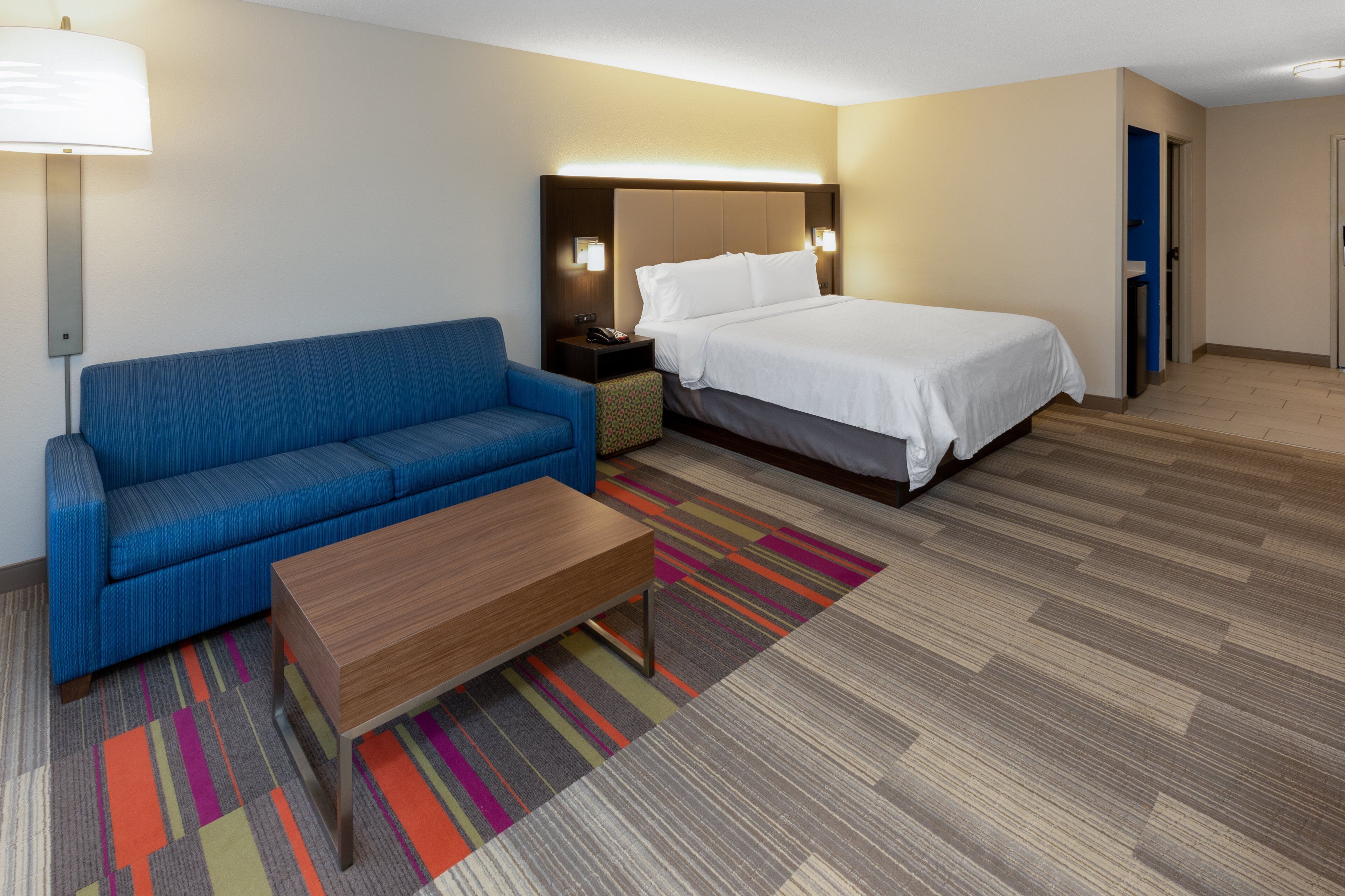 A hotel room with a bed, a blue sofa, a wooden coffee table, and a colorful rug.