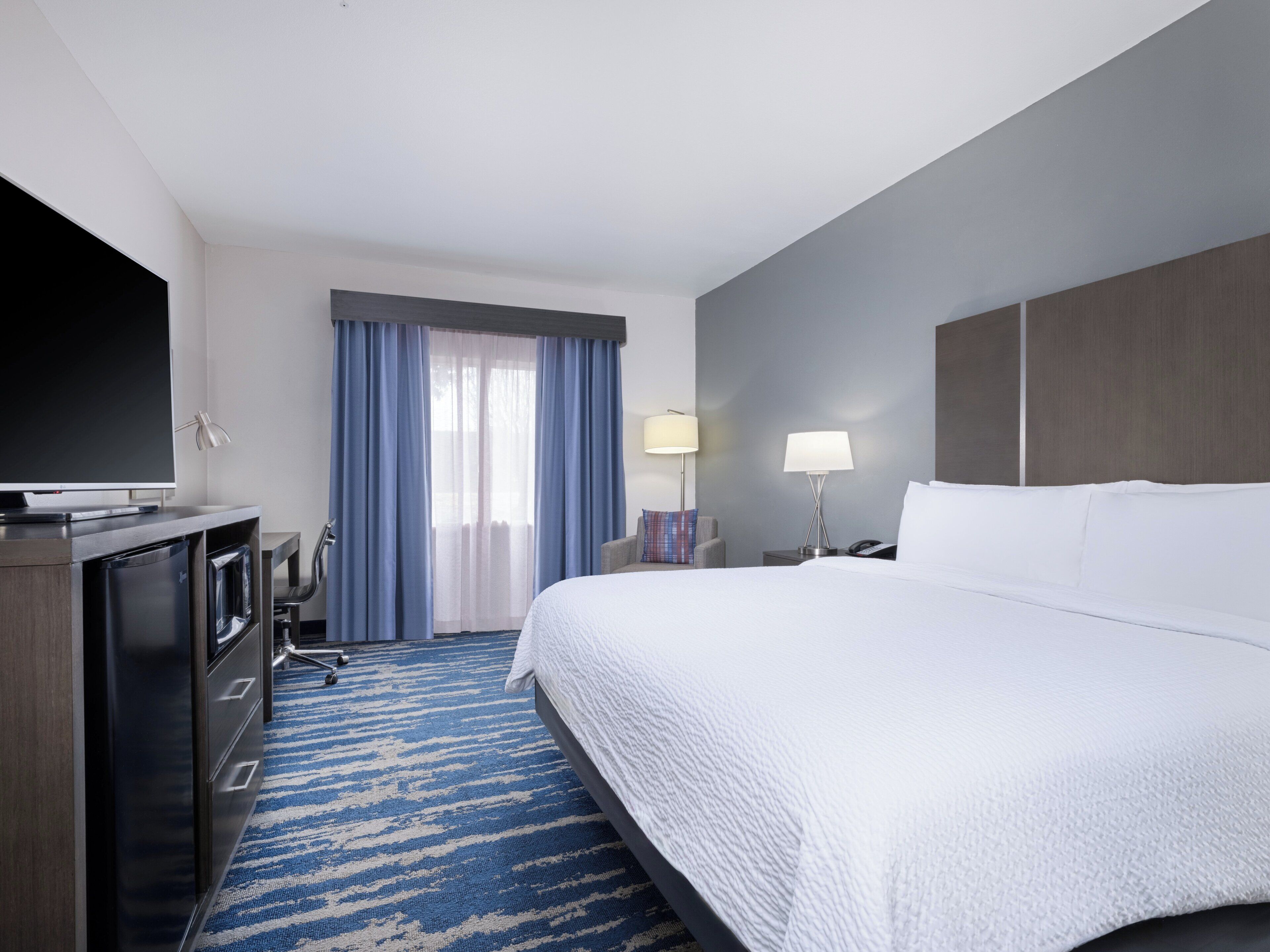 Ein Hotelzimmer mit einem großen Bett, einem Flachbildfernseher, einem Schreibtisch mit Computer, einem Sessel und einem Vorhangfenster.