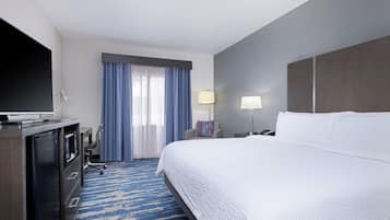 Ein Hotelzimmer mit einem großen Bett, einem Flachbildfernseher, einem Schreibtisch mit Computer, einem Sessel und einem Vorhangfenster.
