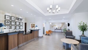 A hotel reception area with a wooden reception desk, framed artwork on the wall, a chandelier, seating area with chairs, and a potted plant.