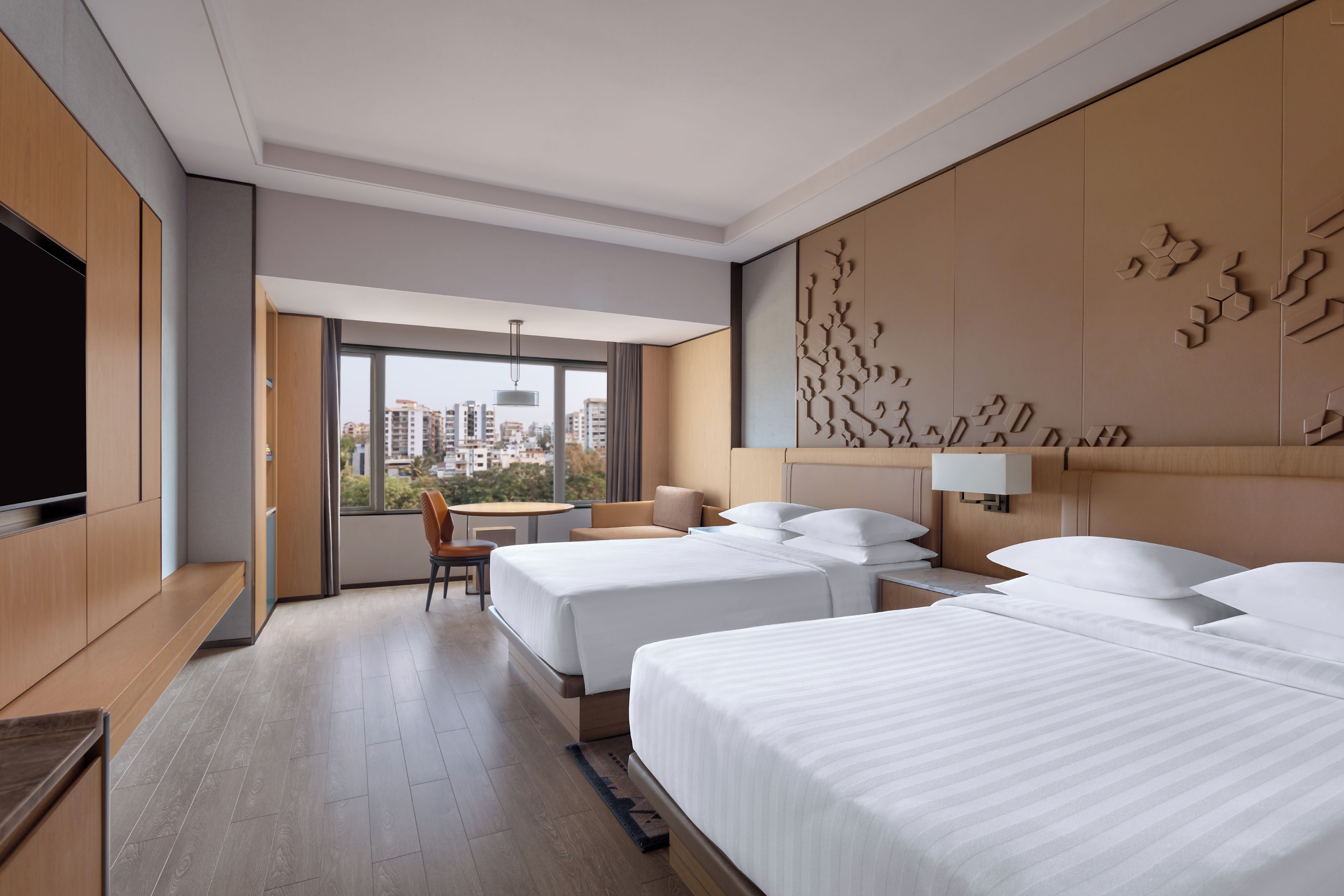 A hotel room with two beds, a TV, a desk, and a view of the city.