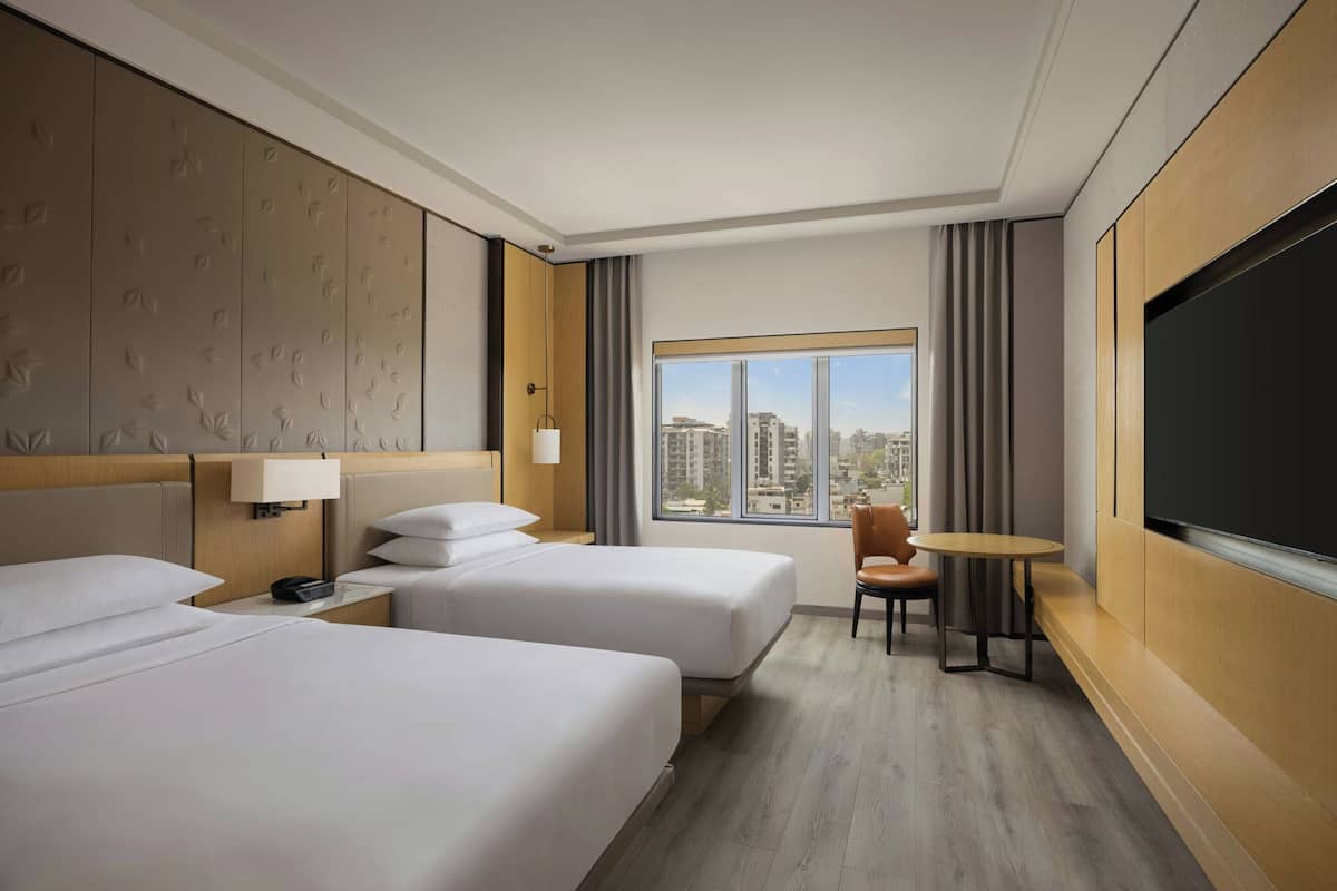Quarto de hotel com duas camas, televisão, uma mesa pequena e janela vista para a cidade.