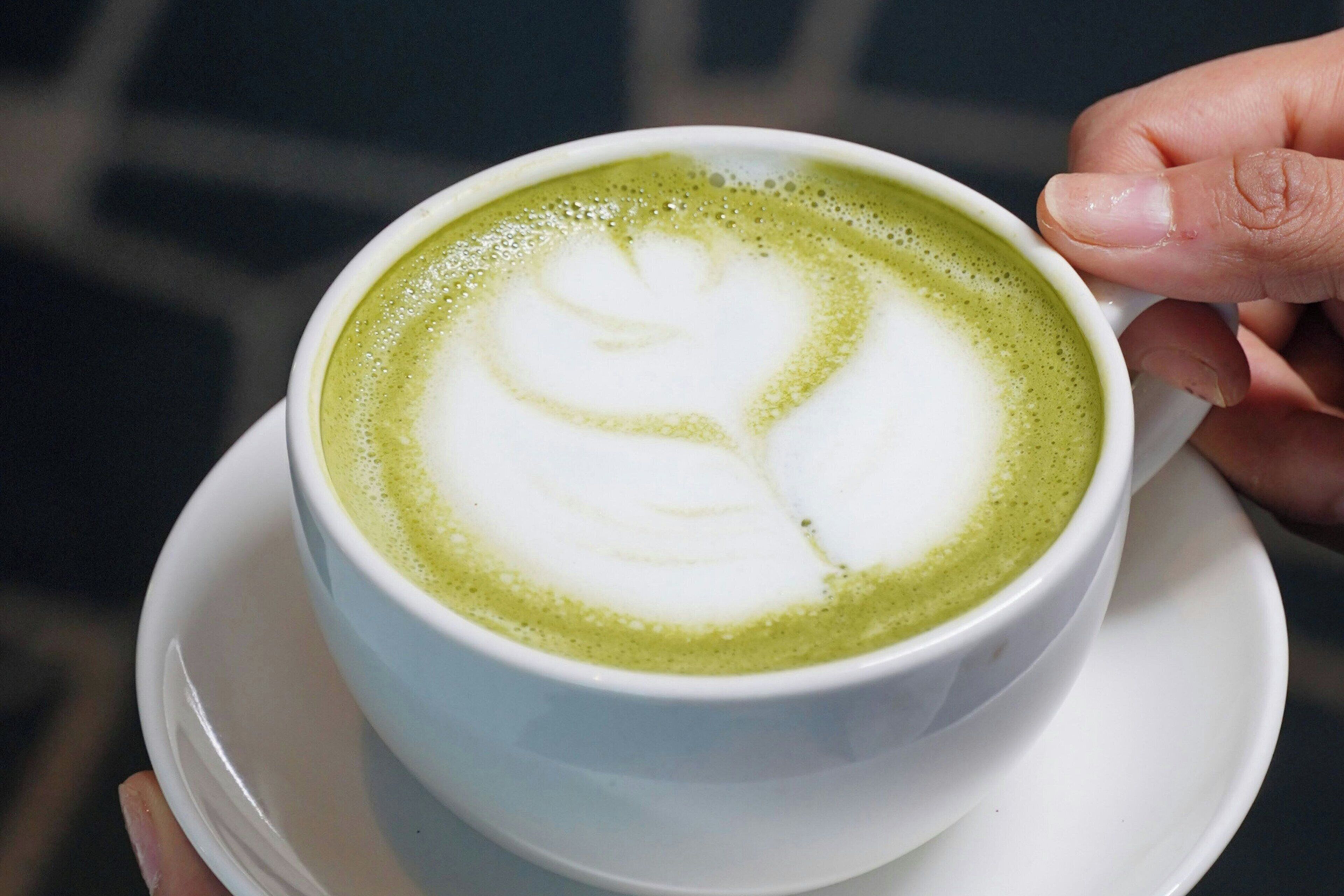En hånd holder en kop grøn te latte med et hjerte tegnet i skummet.