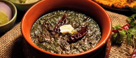 Un tazón de plato verde oscuro y hojas, con una cucharada de mantequilla y chiles rojos encima, rodeado de otros tazones y pan plano.