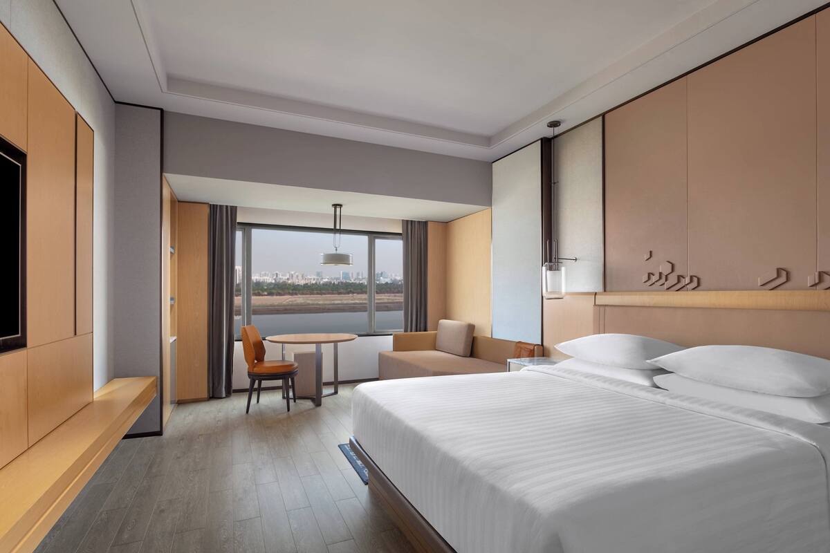 Quarto de hotel moderno com uma cama grande, área de estar com uma mesa e vista da cidade através de grandes janelas.