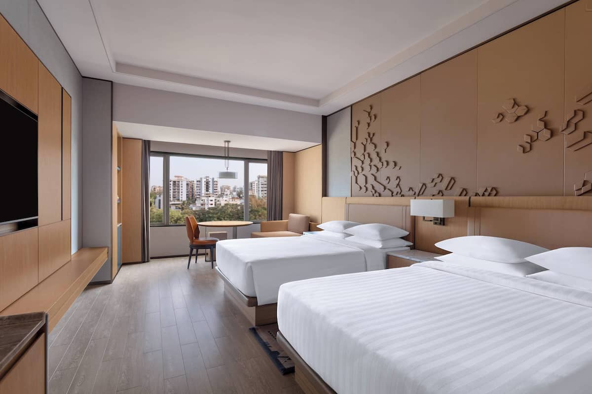 Quarto de hotel moderno com duas camas, televisão, escrivaninha e vista para a paisagem urbana.
