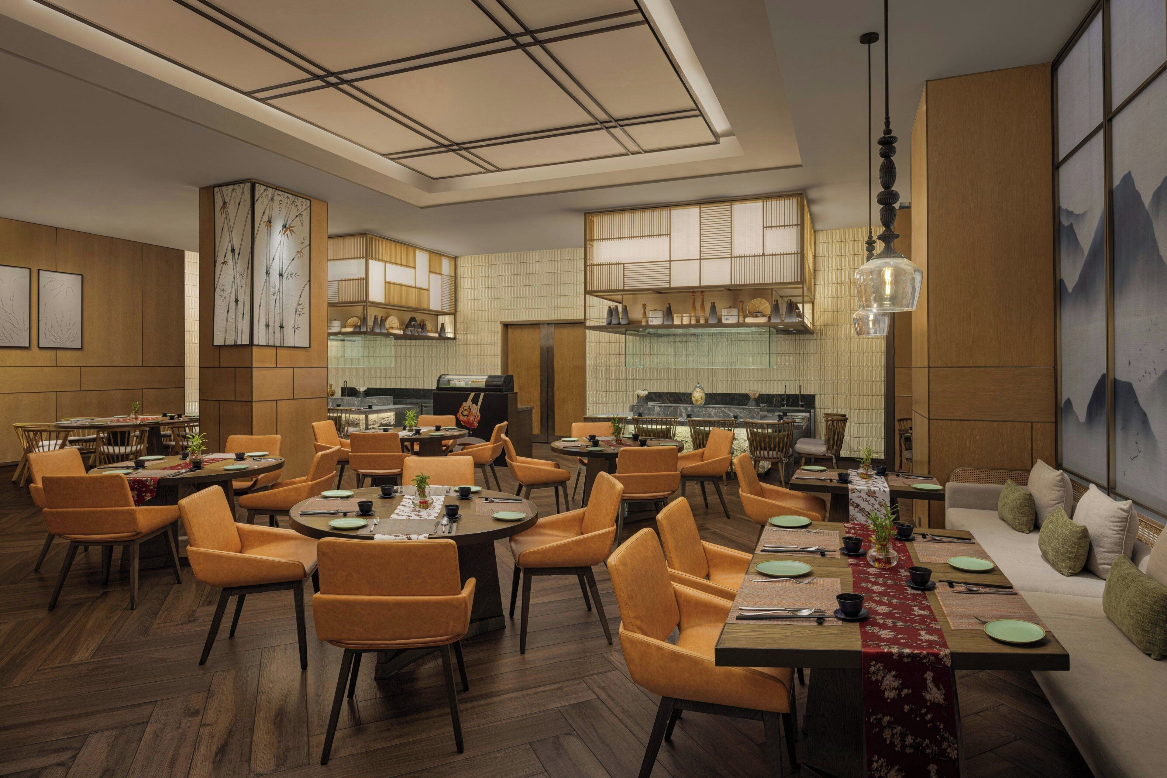 A modern restaurant interior with multiple dining tables, orange chairs, and a kitchen area in the background.
