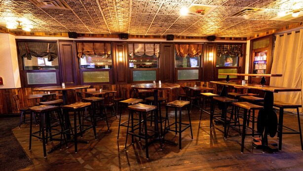 A bar area with wooden tables and stools, a patterned ceiling, and a bar counter with bottles and glasses.