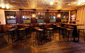 A bar area with wooden tables and stools, a patterned ceiling, and a bar counter with bottles and glasses.