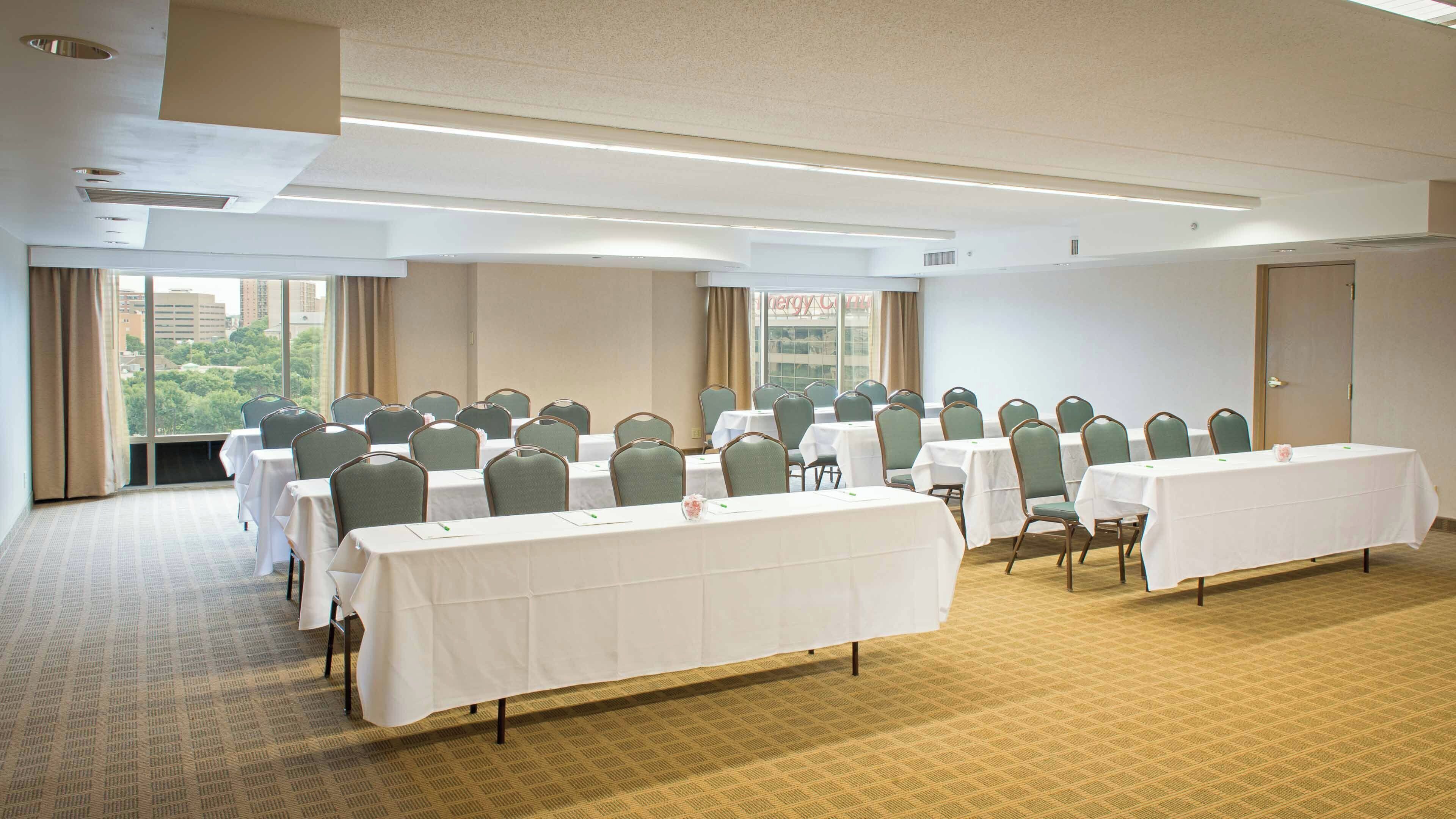 Una sala conferenze con diversi tavoli e sedie, ampie finestre che si affacciano su edifici e alberi, e un pavimento rivestito di moquette.