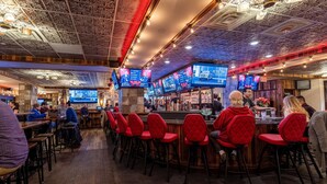 A bar area with multiple TVs displaying ads, red cushioned bar stools, and a wooden floor.