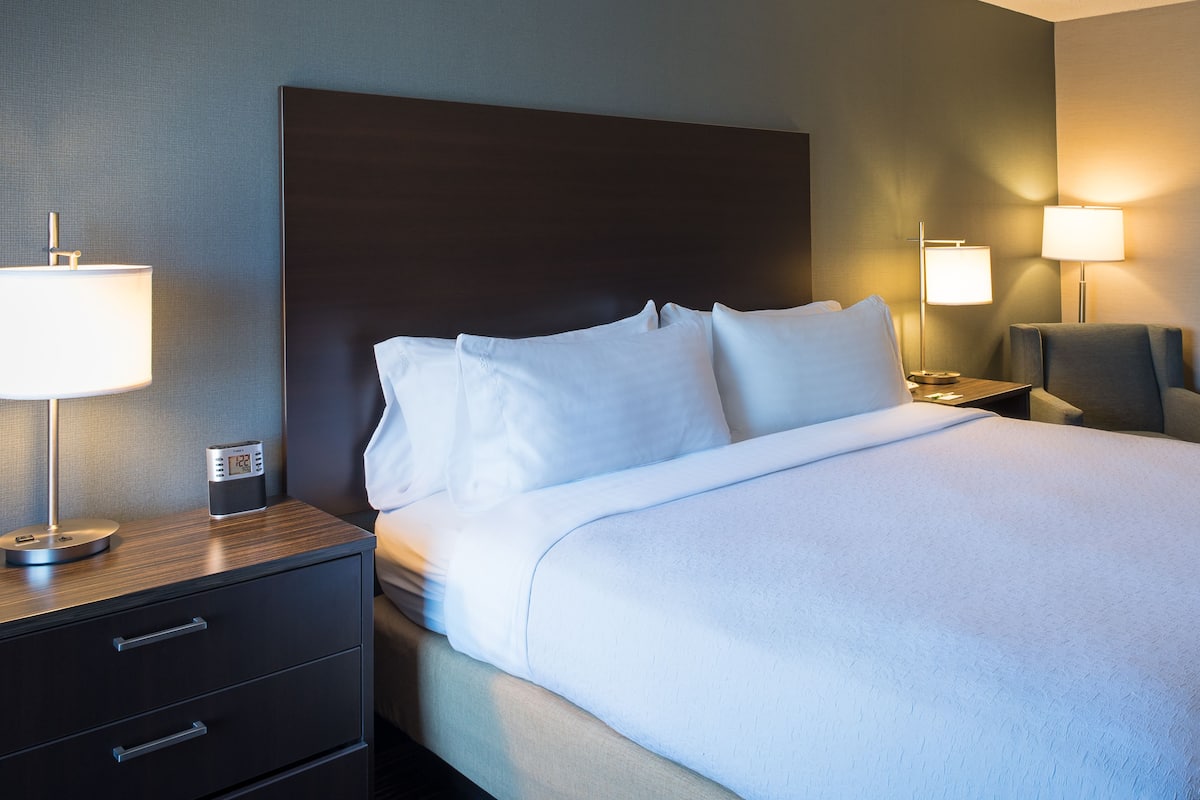 Ein Hotelzimmer mit einem großen Bett, zwei Nachttischen inklusive Lampen, einem Sessel und einem kleinen Tisch.
